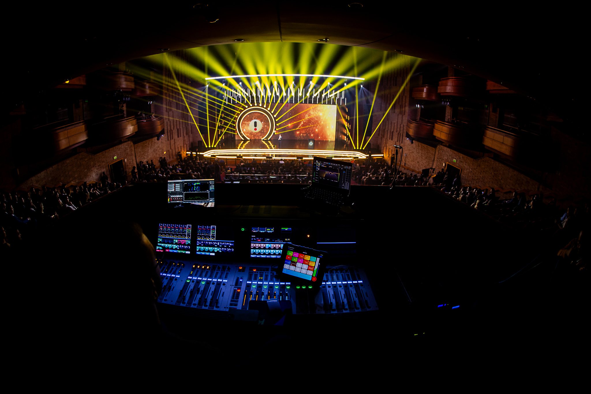 Palco do iBest 2026 visto da mesa de áudio, com feixes de luz amarela